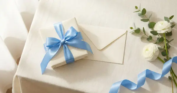 Ivory wedding gift box tied with a soft blue satin ribbon beside a blank cream envelope, white ranunculus, and eucalyptus on a linen tablecloth