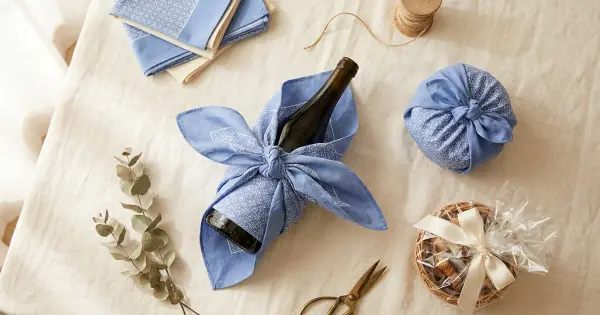 A wine bottle being wrapped in blue furoshiki fabric beside a fabric-wrapped sphere and a cellophane gift basket on a linen table