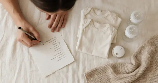 Parent checking items off a baby registry checklist beside organized baby gear - onesies, bottles, and a soft blanket