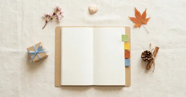 An open planner notebook with four seasonal page tabs surrounded by a spring blossom, seashell, maple leaf, pinecone, and a small kraft gift box with a blue ribbon on warm linen