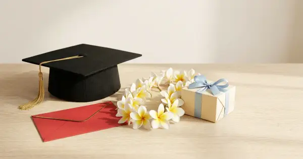 Black graduation cap with a gold tassel beside a red envelope, a white plumeria lei, and a small gift box tied with a blue ribbon on a light wood table