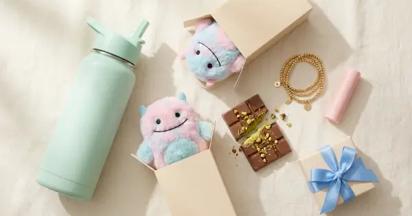 Flat-lay of TikTok-viral gift trends including a pastel water bottle, blind-box plush toys, pistachio chocolate, and a blue-ribbon gift box