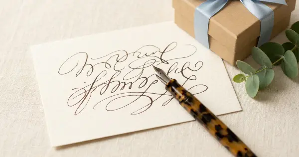 A handwritten thank-you note card with a fountain pen beside a kraft gift box tied with a soft blue ribbon