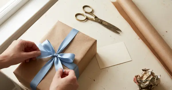 Hands tying a fresh blue ribbon bow on a gift re-wrapped in kraft paper, beside scissors, a blank notecard, and a crumpled piece of old wrapping paper