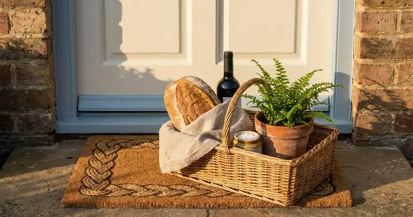 Housewarming gift basket with a rustic loaf of bread, a bottle of wine, a jar of salt, and a potted plant on a doormat at a soft blue front door